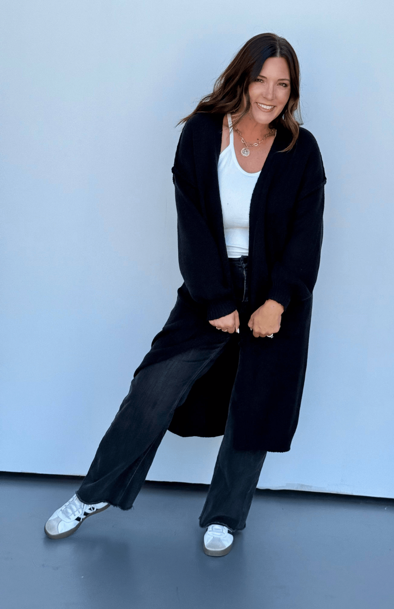 A woman posing in a black long cardigan, white tank top, dark wide-leg jeans, and white sneakers against a light blue studio backdrop.