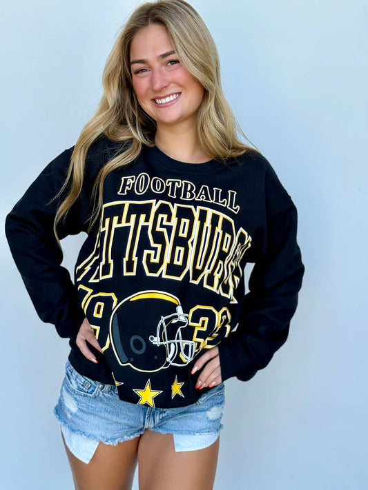 A smiling blonde woman wearing a black long-sleeve sweatshirt with a large 'Pittsburgh 1923 Football' graphic in white and yellow, paired with denim shorts, against a light blue background.