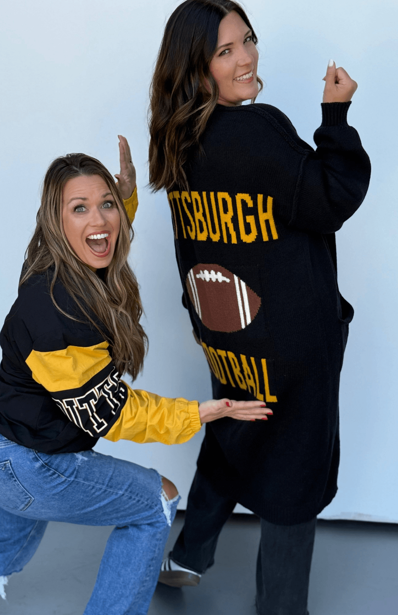 Two women modeling a Pittsburgh football themed sweatshirt. One woman is crouching in the foreground showing the yellow and black sleeve detail, while the other stands behind her showing the back of the oversized black sweatshirt with 'PITTSBURGH FOOTBALL' in yellow varsity lettering and a football graphic.