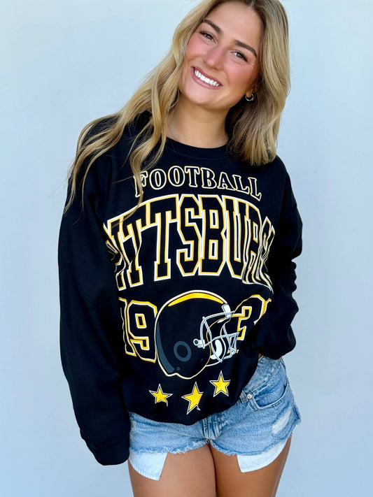 A smiling blonde woman wearing a black long-sleeve sweatshirt with a large gold and white 'Pittsburg 1947 Football' graphic on the front, paired with light-wash denim shorts, against a light blue background.